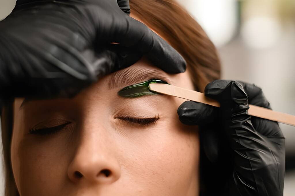 Waxing in Rochester. A close-up of a beauty specialist applying green wax to shape a client’s eyebrows, showcasing the precision and care provided during facial waxing services at a professional laser spa salon.